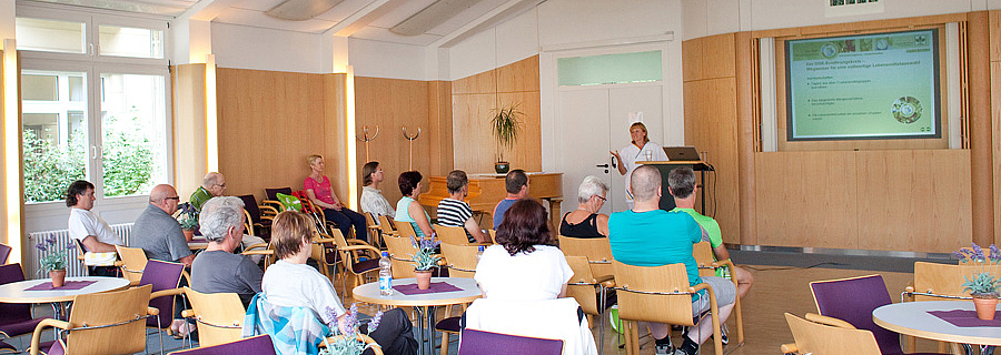 Eine Mitarbeiterin der RehaZentren Baden-Württemberg spricht zu einer Gruppe von Frauen und Männern in einem Schulungsraum.